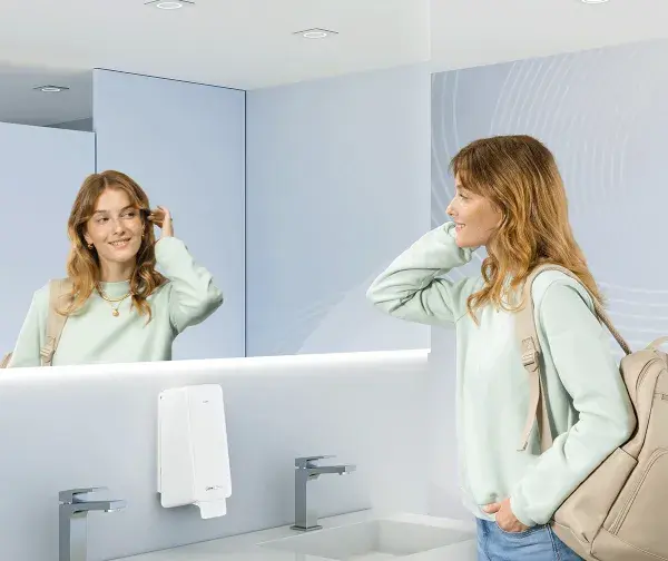 Girl in casual outfit looking at herself in the mirror in a blue washroom equipped with CWS Hygiene PureLine